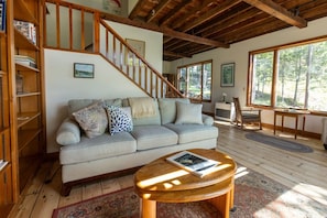 A living room with a sofa, a coffee table, a bookshelf, a staircase, and large windows. - New! Cute boathouse set right on the water (Boothbay)