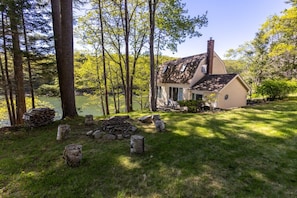 A cabin with a chimney, surrounded by trees and a grassy area with a pile of firewood. - New! Cute boathouse set right on the water (Boothbay)