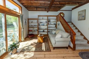 A living room with a wooden staircase, a sofa, a bookshelf, and a large window. - New! Cute boathouse set right on the water (Boothbay)