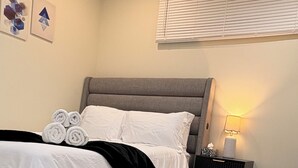 A bedroom with a bed, a bedside table, a lamp, a window with blinds, and a wall with abstract art. - The Dolak Comfort Homes - beautiful, cozy,new, close the airport, grocery stores (Calgary)