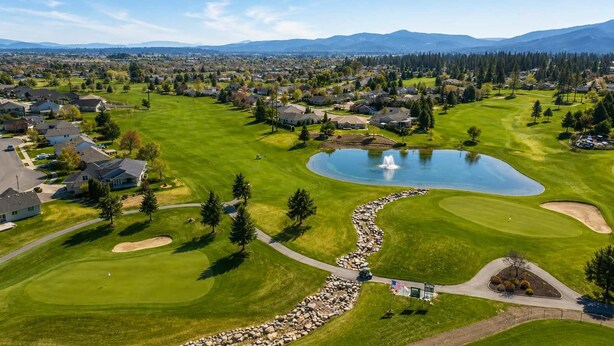 Um campo de golfe com um lago, casas e montanhas ao fundo.