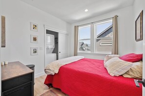 A bedroom with a bed, a desk, a wardrobe, and a window with a view of houses.