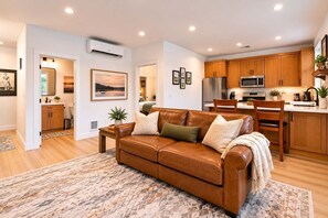 A modern living room with a brown leather sofa, a kitchen with wooden cabinets, and a dining area.
