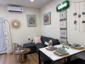 Una sala de estar con sofá, una mesa de centro, una mesa de comedor con individuales y platos, aire acondicionado instalado en la pared, una obra de arte floral enmarcada y un letrero de neón que dice 'Welcome'.