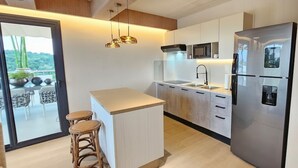 A modern kitchen with a large refrigerator, a microwave, and a sink. There is a small island with stools and a view of the outdoors through a glass door. - MŪTE Collection at Spirit of Terrenas (Las Terrenas)