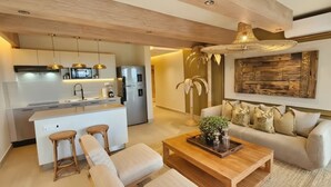 A modern kitchen with white cabinetry, stainless steel appliances, and pendant lighting. There is a dining area with a wooden table and a sofa with cushions. - MŪTE Collection at Spirit of Terrenas (Las Terrenas)