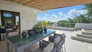 An outdoor patio with a glass table, wicker furniture, potted plants, and a view of the ocean. - MŪTE Collection at Spirit of Terrenas (Las Terrenas)