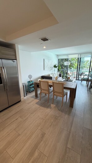 Een moderne keuken met een roestvrijstalen koelkast, een houten eettafel met stoelen en uitzicht op de buitenlucht door grote ramen.