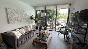 Een moderne woonkamer met een grijze bank, een bijzettafeltje met boeken en kaarsen, en uitzicht op een terras met stoelen en een tafel.