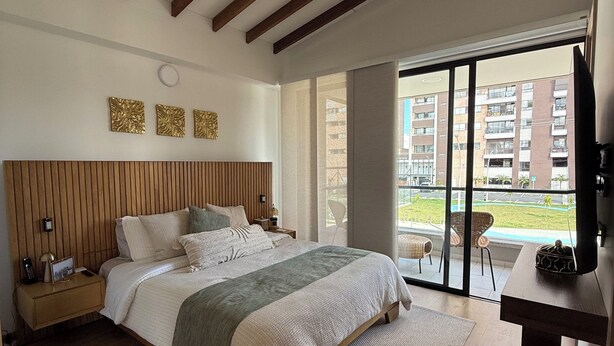 Une chambre à coucher comprenant un lit, des tables de chevet, un bureau, une télévision et une grande fenêtre donnant sur des immeubles.