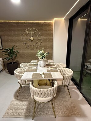 Un comedor con pared de ladrillo, un reloj de pared grande, un jarrón con flores y un ventanal de vidrio.