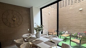 Un espace de repas moderne, agrémenté d’une grande horloge, d’un vase de fleurs et offrant une vue sur un mur de briques et sur la verdure à l’extérieur.