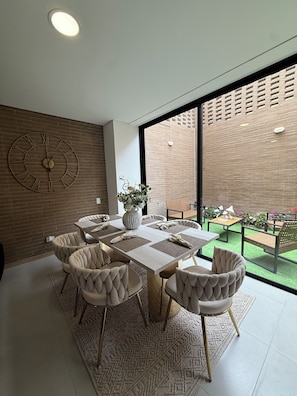 Un comedor moderno con un reloj grande, un florero con flores y vistas a un muro de ladrillo y follaje exterior.