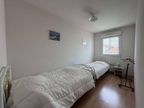 Ein Schlafzimmer mit zwei Einzelbetten, einem Fenster mit Jalousien, einem Heizkörper und einem kleinen Nachttisch mit Lampe.