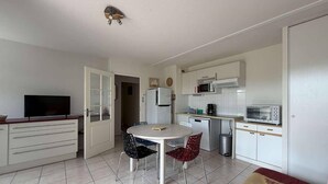 Cozinha compacta equipada com micro-ondas, torradeira e frigorífico. Inclui uma mesa de jantar com duas cadeiras e uma televisão sobre um móvel.