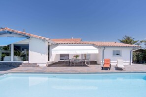 Un espace aménagé au bord de la piscine, avec une table et des chaises, une terrasse couverte et un ciel d’un bleu éclatant.