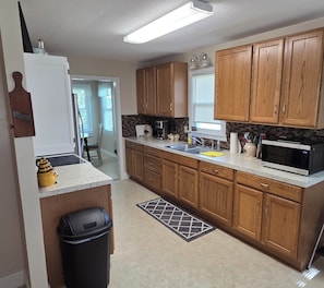 A kitchen with wooden cabinets, a stainless steel sink, a microwave, and a trash bin.