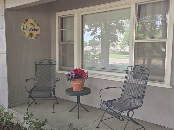 A porch with two metal chairs, a small table, a welcome sign, and a window with blinds.