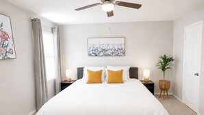 A bedroom with a bed, a bench, a ceiling fan, a nightstand, a lamp, a plant, and a door. - [GS] 3BR+3BR I 2 Kitchens I Two Adjacent Townhomes I Brand New & Family-Friendly (San Antonio)