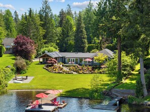 Uma casa com gramado bem cuidado, um píer com cadeiras e guarda-sóis, e um lago.