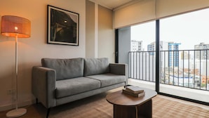 Ein modernes Wohnzimmer mit einem grauen Sofa, einer Stehlampe, einem runden Holztisch und einem großen Fenster mit Blick auf Gebäude.