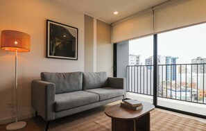 A modern living room with a grey sofa, a floor lamp, a round wooden coffee table, and a large window with a view of buildings. - w* | Upscale 2BR w/ Balcony & View in Miraflores (Miraflores)