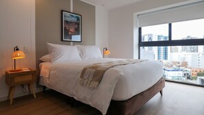Ein modernes Hotelzimmer mit einem großen Bett, zwei Nachttischlampen, Blick auf die Stadt und einem gerahmten Bild an der Wand.