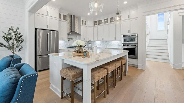 Una cocina moderna con armarios blancos, una isla central con taburetes, electrodomésticos de acero inoxidable y una lámpara colgante.