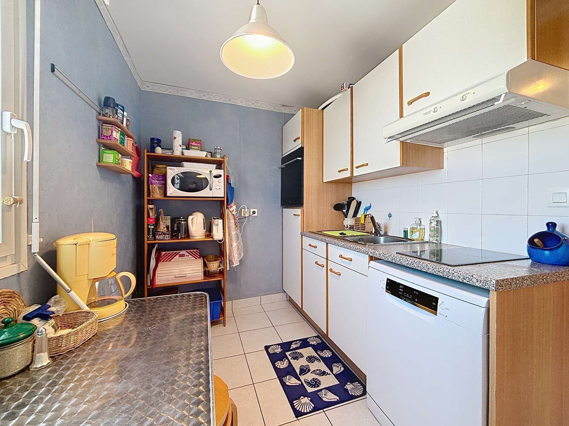A compact kitchen with a microwave, stove, and dishwasher. A wooden shelf holds various kitchen items.