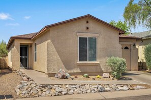 A single-story house with a beige stucco exterior, a front door, a window with shutters, and a small garden with rocks and cacti. - Northwest Tucson Retreat | BBQ & Family-Friendly (Tucson)
