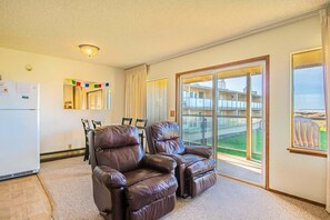 A living room with two leather recliners, a dining area with chairs, a refrigerator, and a large window with a view of buildings.