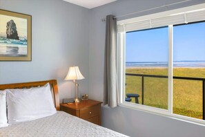 A bedroom with a bed, a nightstand, a lamp, a framed picture of a rock formation, and a window with a view of a grassy field. - Hi-Tide Resort, Top Floor, Queen Bed, Unit 32 (Moclips)