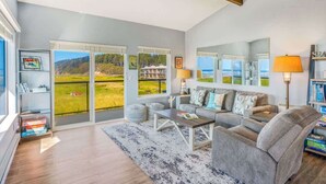 A spacious living room with a large window, a sofa set, a coffee table, and a bookshelf.