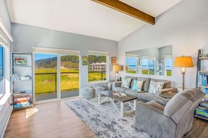 A spacious living room with a large window, a sofa set, a coffee table, and a bookshelf.