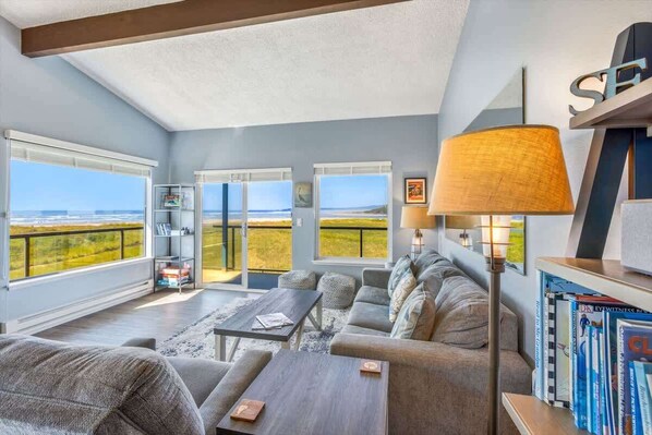 A modern living room with a sofa, coffee table, and bookshelves. Large windows offer a view of the landscape. - Hi-Tide Resort, Top Floor, Queen Bed, Unit 32 (Moclips)