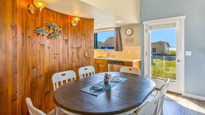 A dining area with a round table, white chairs, wooden paneling, a kitchen in the background, and a view through a glass door.