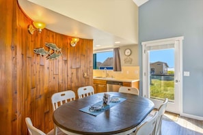 A dining area with a round table, white chairs, wooden paneling, a kitchen in the background, and a view through a glass door.