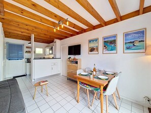 A modern kitchen with white cabinets, a dining area with a wooden table and chairs, and a TV mounted on the wall. - Erdeven - 3-room house - 42m² - Wifi - 1* (ERDEVEN)