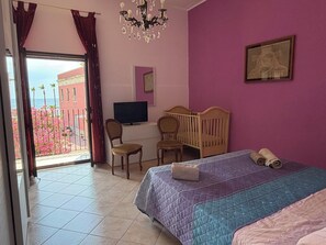A bedroom with a bed, a desk, a chair, a crib, a chandelier, and a window with curtains. - Sea view terrace (Siracusa)