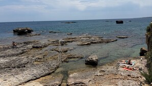 Costa rocosa con agua cristalina, algunas personas tomando el sol y tierra a lo lejos.