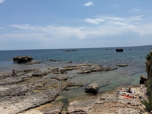 A rocky shoreline with clear water, a few people sunbathing, and distant land. - Sea view terrace (Siracusa)
