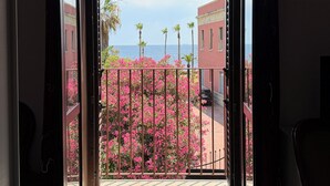 Puerta abierta que da a un balcón con vistas a un arbusto de flores rosadas, palmeras y edificios al fondo.