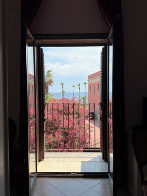 An open door leading to a balcony with a view of a pink flowering bush, palm trees, and buildings in the background. - Sea view terrace (Siracusa)