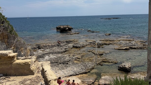 Una costa rocosa con agua cristalina, un velero a lo lejos y una persona sentada sobre una roca.