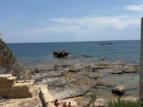 A rocky shoreline with clear water, a distant sailboat, and a person sitting on a rock. - Sea view terrace (Siracusa)