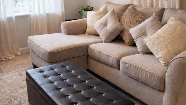 Un canapé d’angle beige agrémenté de plusieurs coussins, un pouf marron foncé et des rideaux en voile léger.