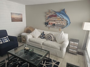 Ein Wohnzimmer mit einem weißen Sofa, einem Glastisch, einem blauen Sessel und einem Wandbild mit einem blauen Fisch.