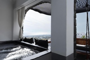 A room with a view of a cityscape, a bathtub, and a window with curtains. - Lumera Sunrise Suite | Heated Jacuzzi & Sea View (Thira)