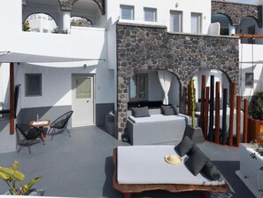 A modern outdoor living area with a stone wall, a white sofa, and a wooden table. - Lumera Sunrise Suite | Heated Jacuzzi & Sea View (Thira)