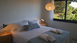 Una cama bien tendida con almohadas, una lámpara de noche y toallas sobre una mesita de madera.